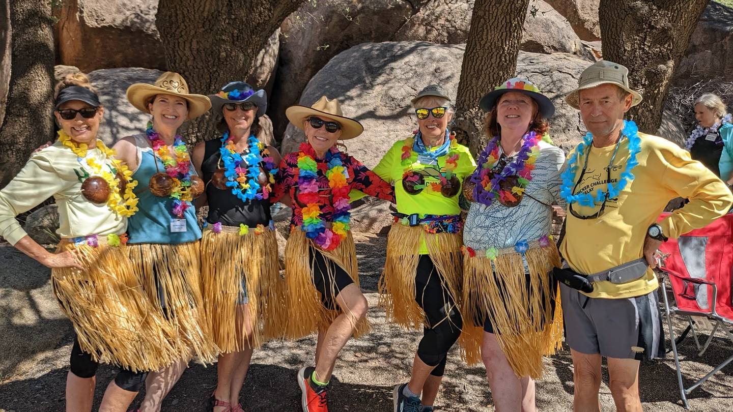 Seven campers in grass skirts, leis, and cowboy hats laughing together in the shade of oak trees