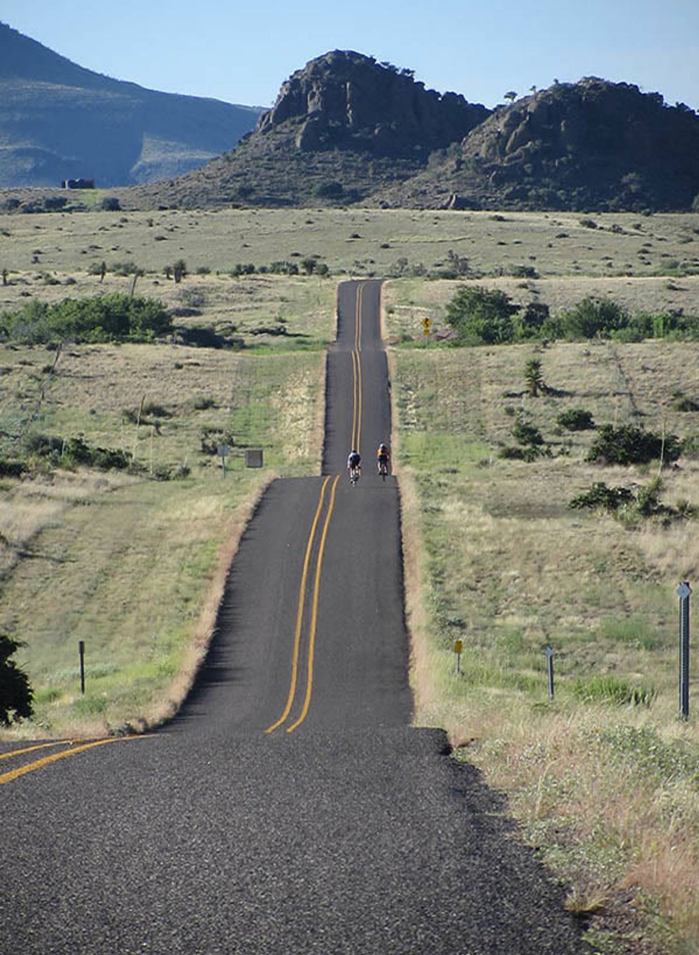 Two cyclists riding a rolling ribbon of empty road toward rocky desert peaks